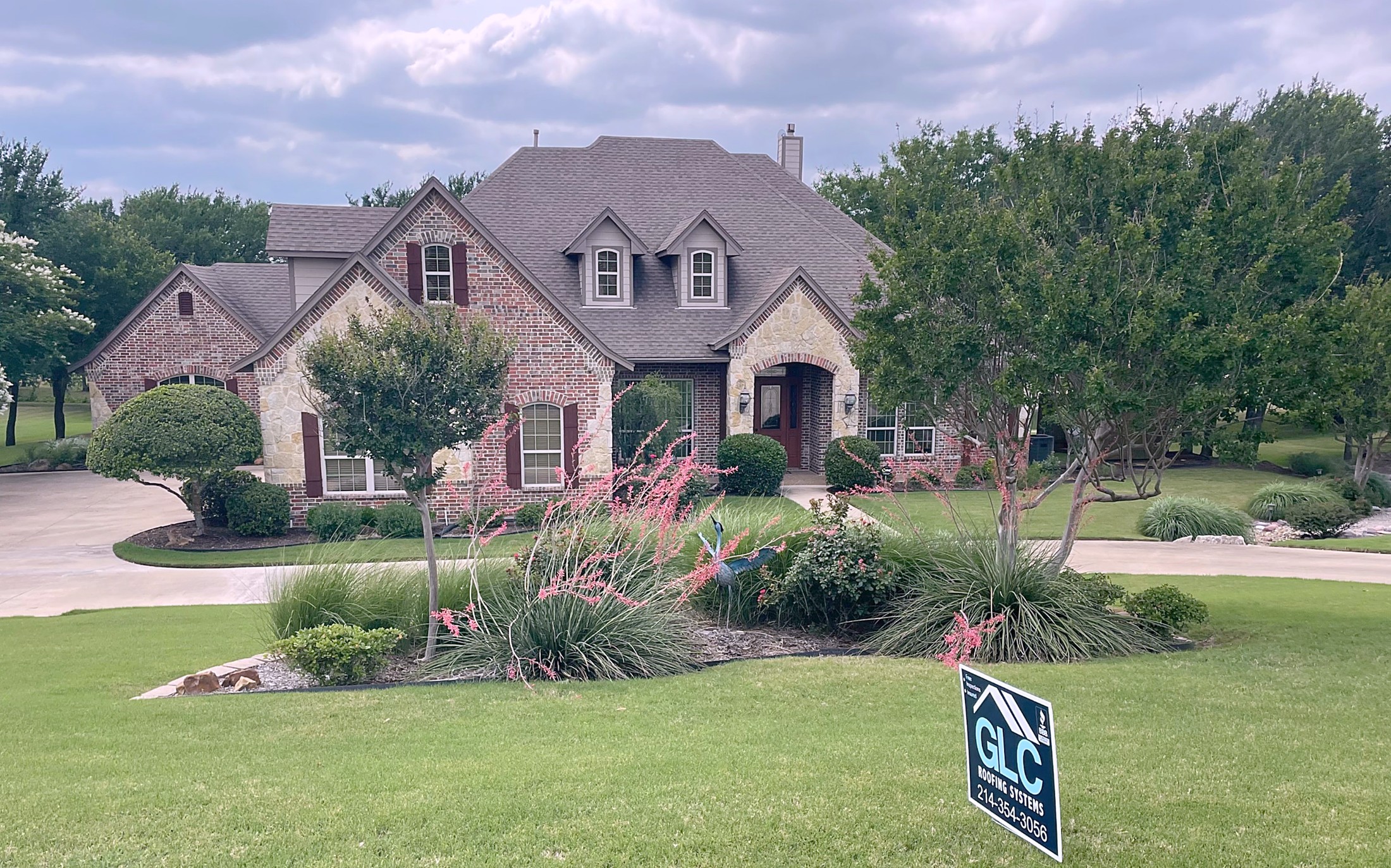 A brick and stone two-story home with a new shingle roof and the GLC Roofing Systems sign on the front lawn